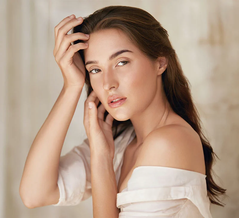 Woman with dark hair and bare shoulders gently touching her face, wearing an off-the-shoulder white top - ClearSilk Laser Treatment in Red Bank, NJ