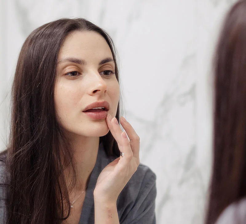 A woman looks into the mirror, touching her lips, with a thoughtful expression - Hylenex in Red Bank, NJ