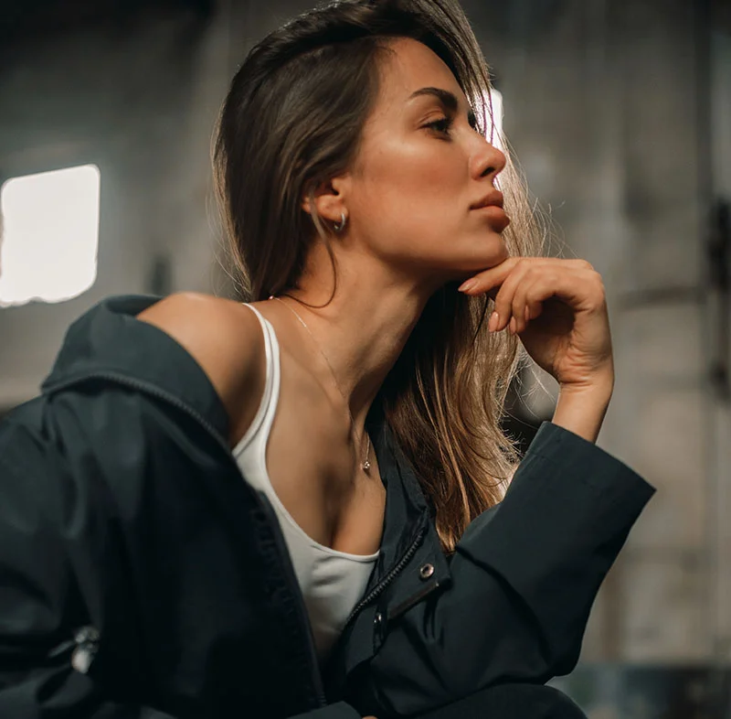 A thoughtful woman with long dark hair, wearing a dark jacket and white top, sitting in a dimly lit industrial space, her hand resting under her chin - Kybella in Red Bank, NJ