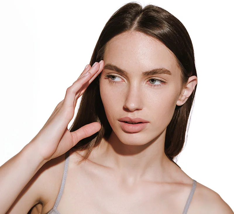 A brunette woman in a grey tank top, gazing to the side with her hand touching her temple - PRP Facial in Red Bank, NJ