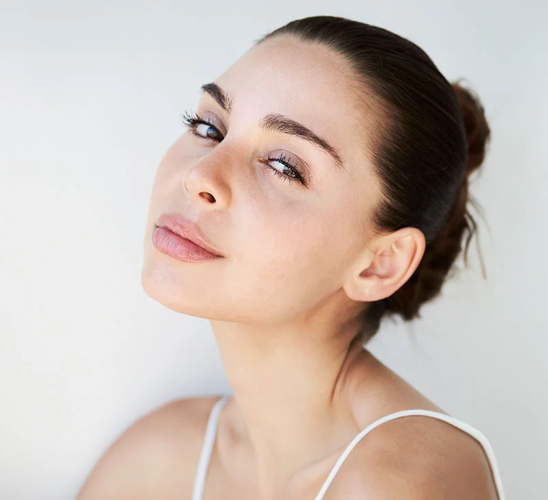 A brunette woman with her hair in a bun, wearing a white tank top with her head slightly tilted back - Rosacea Treatment in Red Bank, NJ